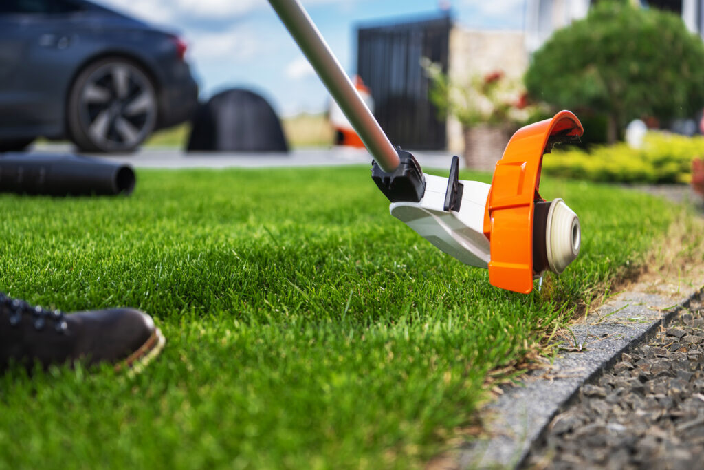 Close-up of an orange lawn mower blade on a grassy lawn with a blurred car and garden in the background.