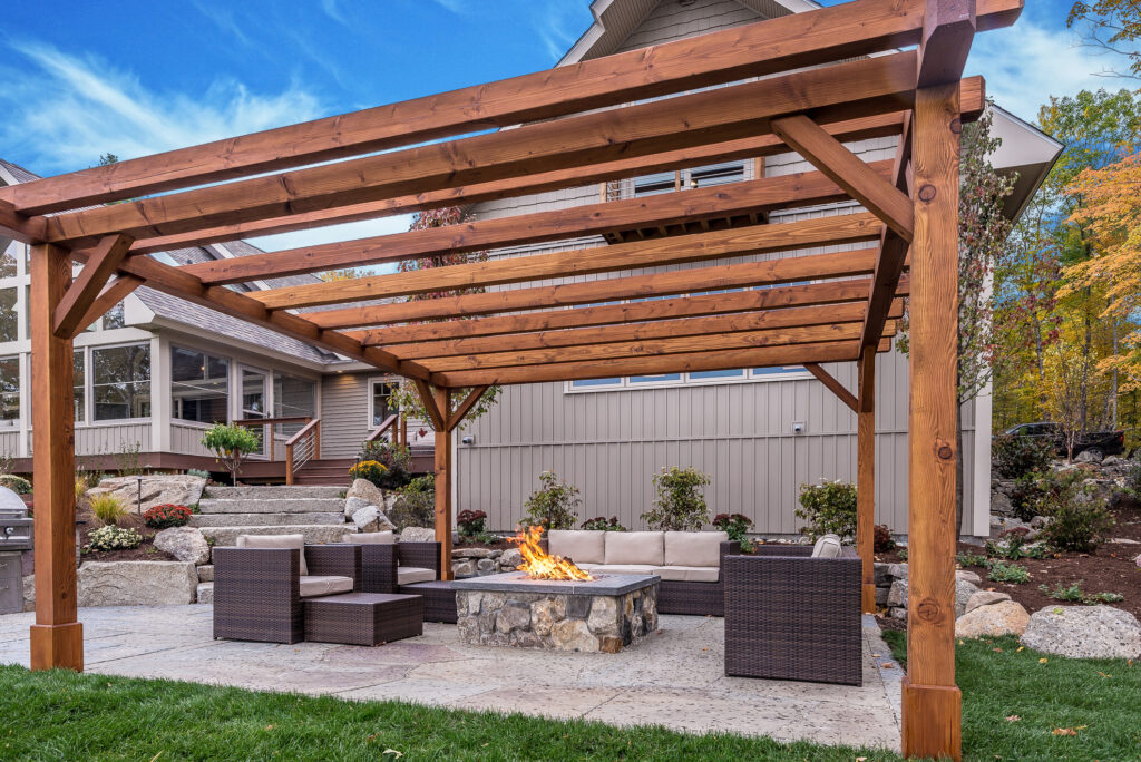 Backyard scene with a wooden pergola, woven seating around a stone fire pit, and a beige house in the background