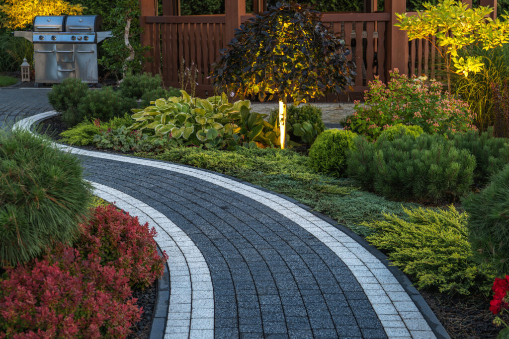 Curved black-and-white paver driveway winding through a lush garden with a stainless grill on the left and a wooden fence in the background.