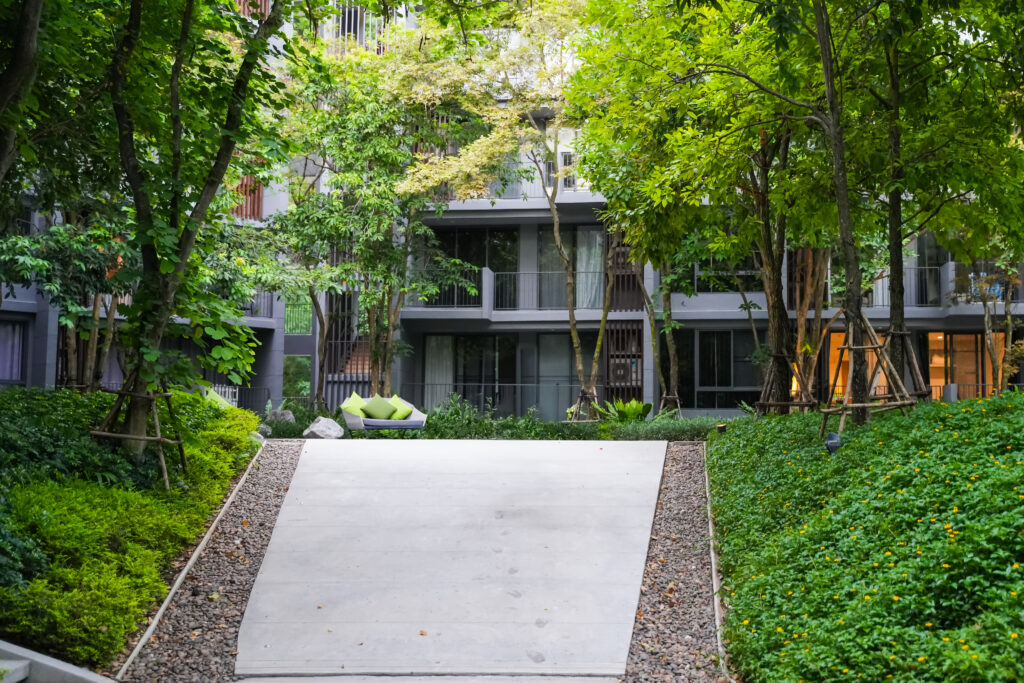 Courtyard with green trees and shrubs, a concrete path leading to a modern apartment building in the background.