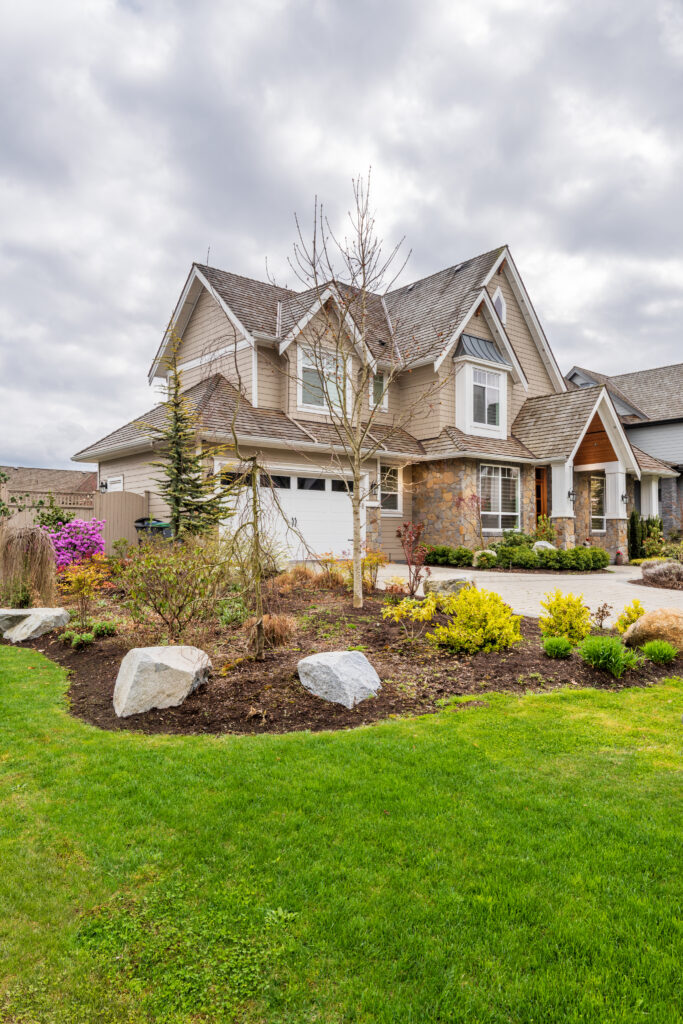 Two-story beige house with a gabled roof, attached garage, and a landscaped front yard with mulch, rocks, and small trees under a cloudy sky.