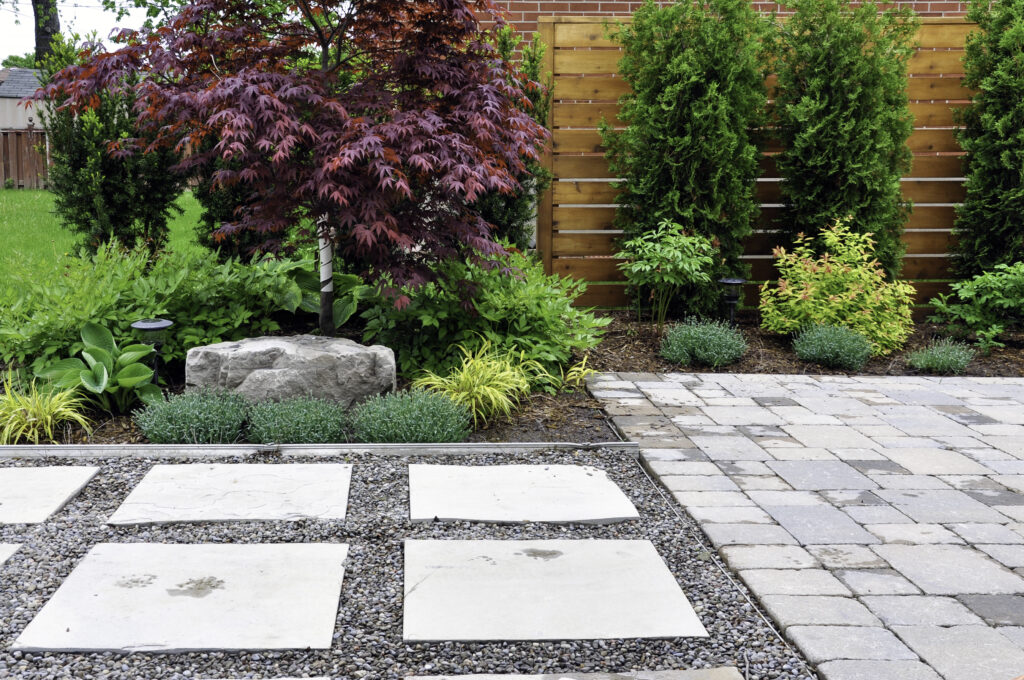 Garden scene with square stepping stones set in gravel, bordered by lush greenery and a red-leaved tree beside a wooden fence.